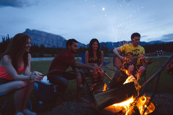 A group of people sits around the campfire at night, one plays the guitar