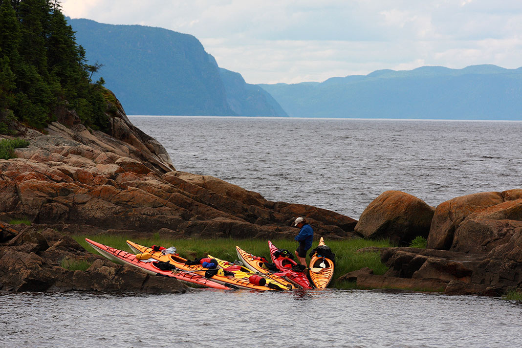 A woman stands near a line of kayaks on the shore in Saguenay