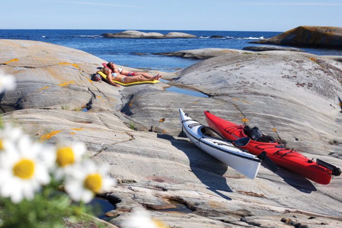 Two women sunbathing on thermarests on rocks with sea kayaks pulled up on shore.