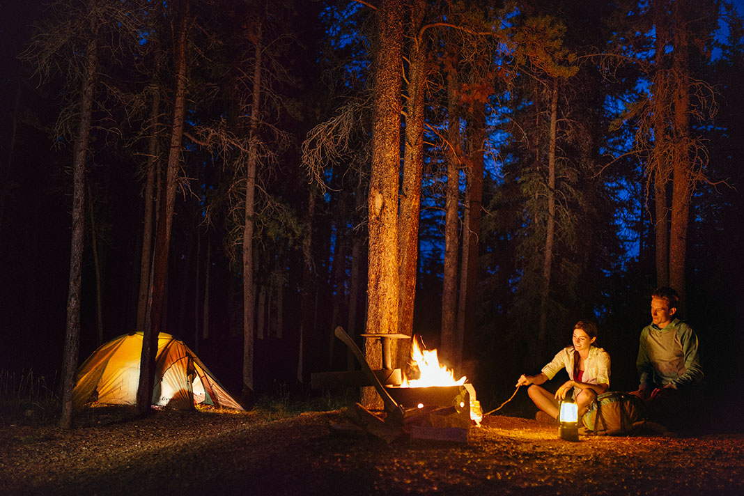 Two people sitting beside campfire in the woods with tent in background