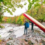 9 Non-Essentials You Must Bring On Your Next Canoe Trip Two people standing at top of waterfall, one with canoe and other with blue barrel.