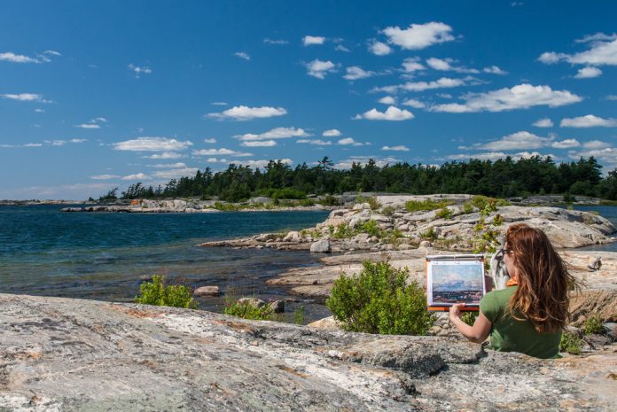 Woman sitting on rocky coast by lake holding painting of same view.