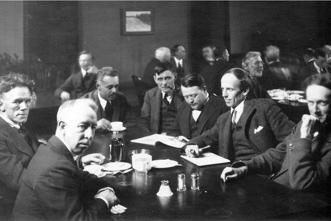 Black and white photo of seven men sitting around a table wearing suits.