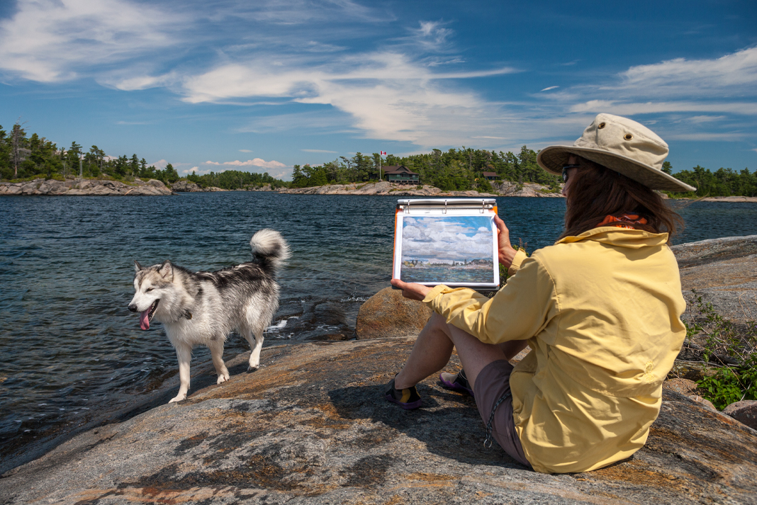 Husky dog walking beside lakeshore, woman sits in foreground holding up painting of same location.