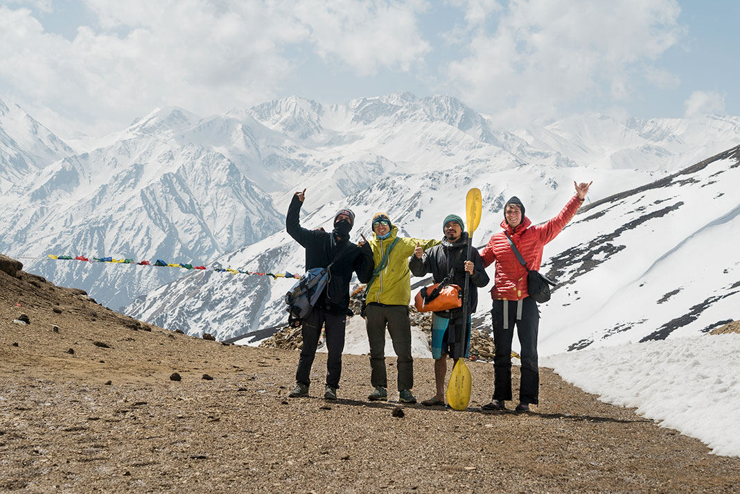 Four people standing with bags and a kayak paddle, with snow-covered mountains in background.