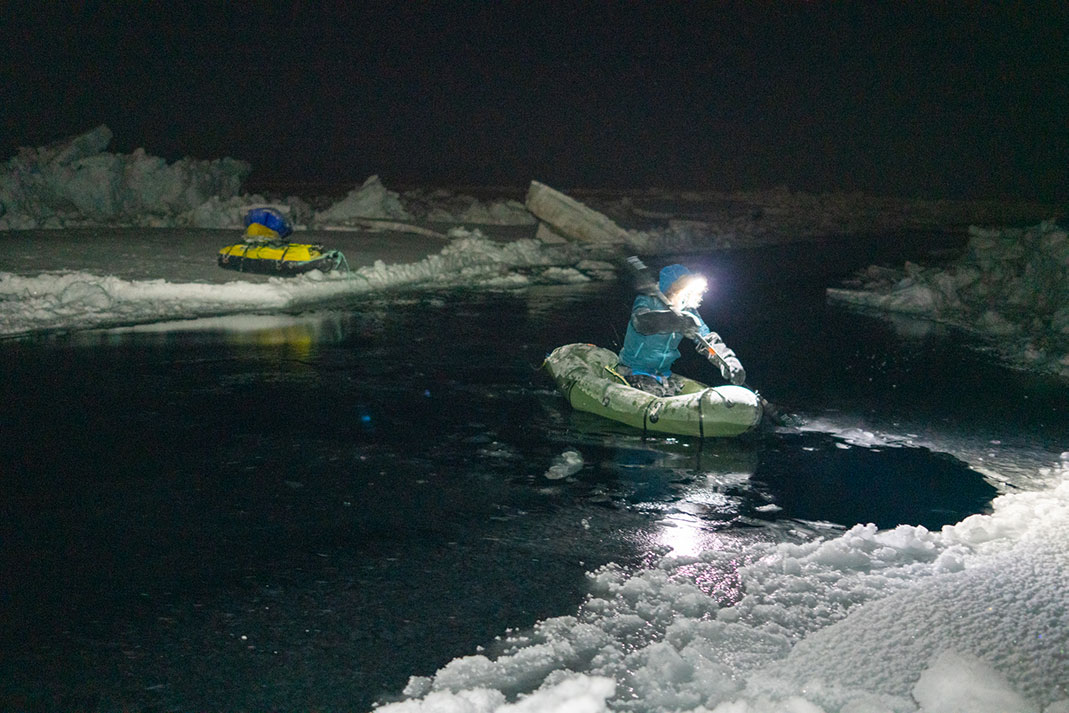 Man paddling small packraft across water between two slabs of ice, with supplies trailing behind.