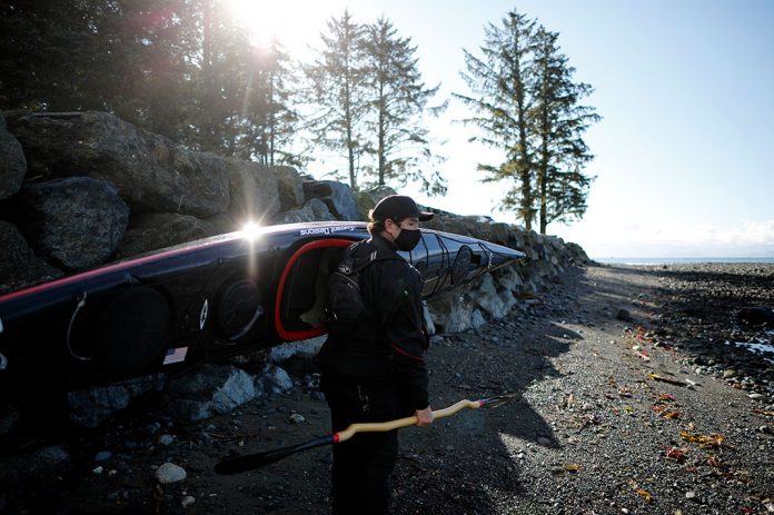 Man wearing a mask and baseball hat carrying a kayak and paddle along the coast.