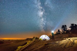 Person standing beside tent and kayak pulled up on shore, with headlight on, and Milky Way overhead.