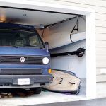 7 Creative & Simple Kayak Storage Ideas Paddleboards leaning up against wall and kayak hanging from ceiling against wall, squeezed beside a van in a garage.