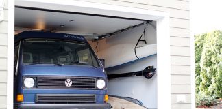 Paddleboards leaning up against wall and kayak hanging from ceiling against wall, squeezed beside a van in a garage.