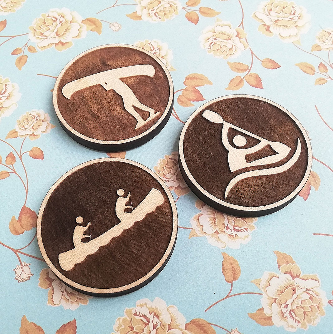 Wooden fridge magnets with person portaging canoe, two people paddling canoe, and person paddling a kayak etched on.