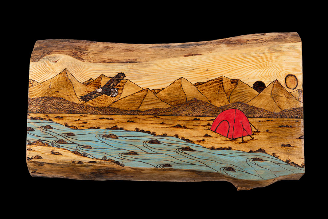Piece of wood with scene of tent beside a river with eagle flying overhead and mountains in background. 