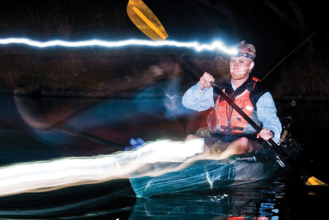 man paddles a kayak at night with headlamp and lights leaving trails