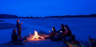 Five people sitting around campfire with sea kayaks pulled up on shore and river in background.