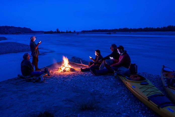 Five people sitting around campfire with sea kayaks pulled up on shore and river in background.