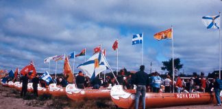 Voyageur canoes waiting on shore amid crowd of people and provincial flags.