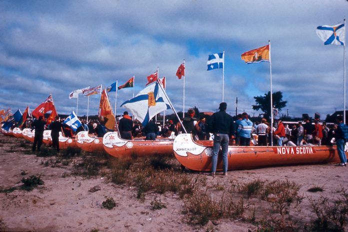 Voyageur canoes waiting on shore amid crowd of people and provincial flags.