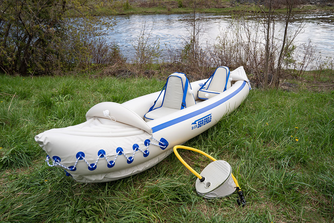 White and blue, tandem inflatable kayak with foot pump sitting on grass beside river.