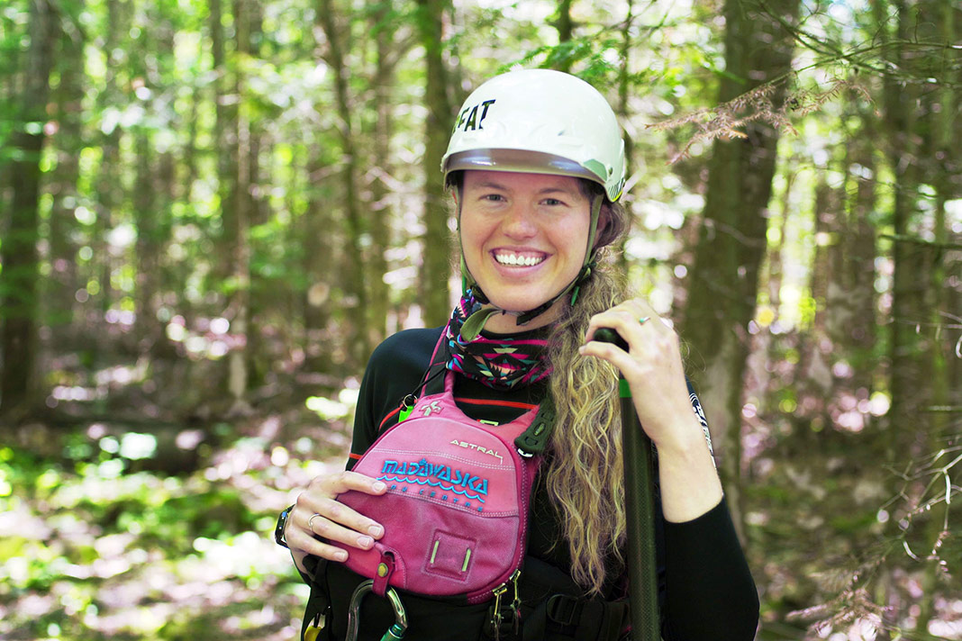 Headshot of woman wearing helmet and PFD, standing in woods.