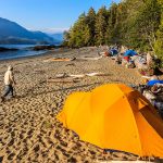 Tents set up on a beach with sea kayaks pulled up on shore, and ocean and mountain in background.