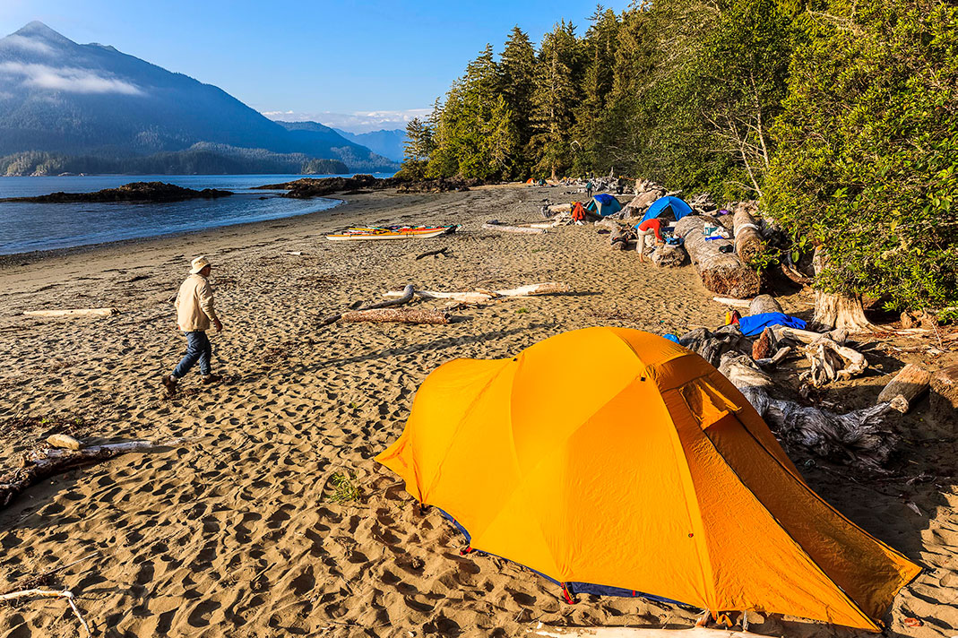 Tents set up on a beach with sea kayaks pulled up on shore, and ocean and mountain in background.
