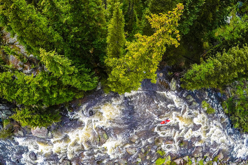 Overhead shot of person in red whitewater kayak on a river with trees along the bank.