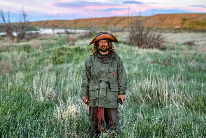 Man wearing large-brimmed hat, dirty coat standing in field with river in background.