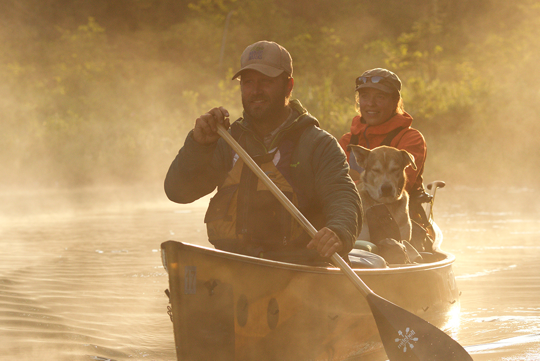 Man and woman paddling canoe, with dog between them.