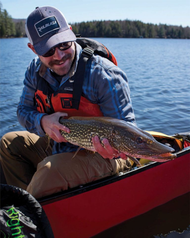 smiling man holds up a northern pike caught from his canoe