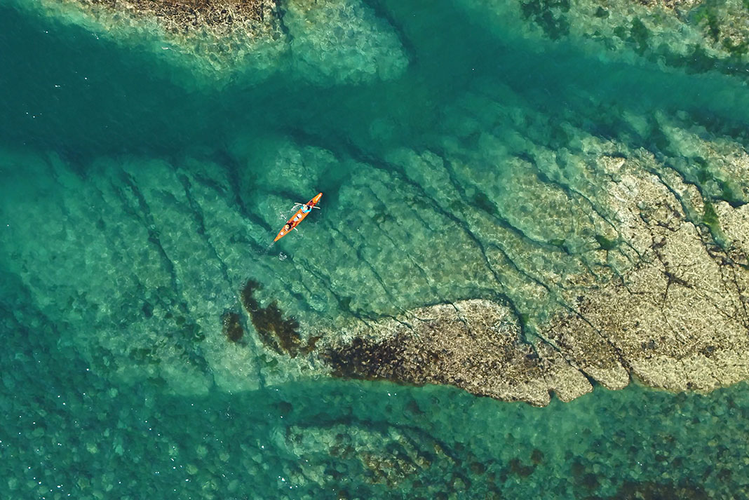 Bird's eye view of orange tandem kayak paddling across clear, turquoise water.