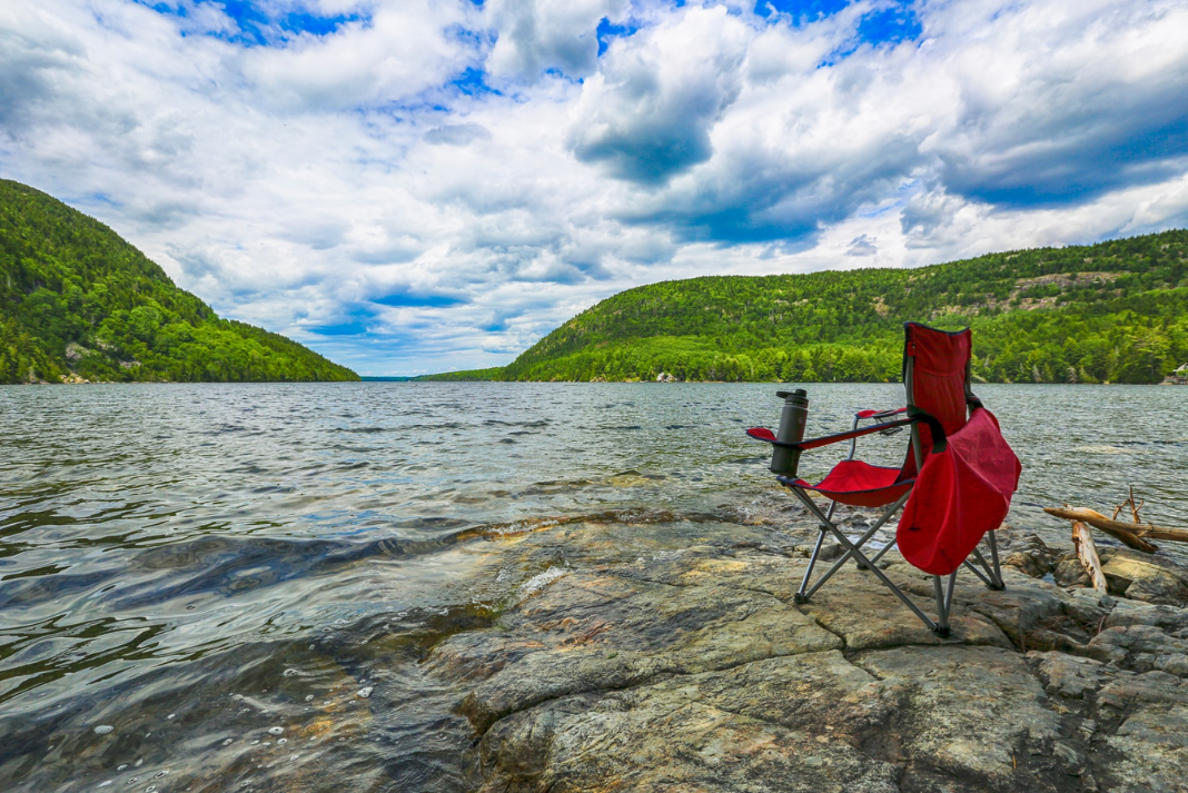 A red camp chair is stationed on a rock overlooking a picturesque lake.