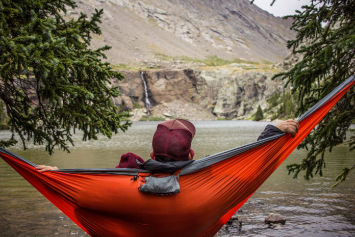 A man lounges in his hammock overlooking a a lake surrounded with mountains