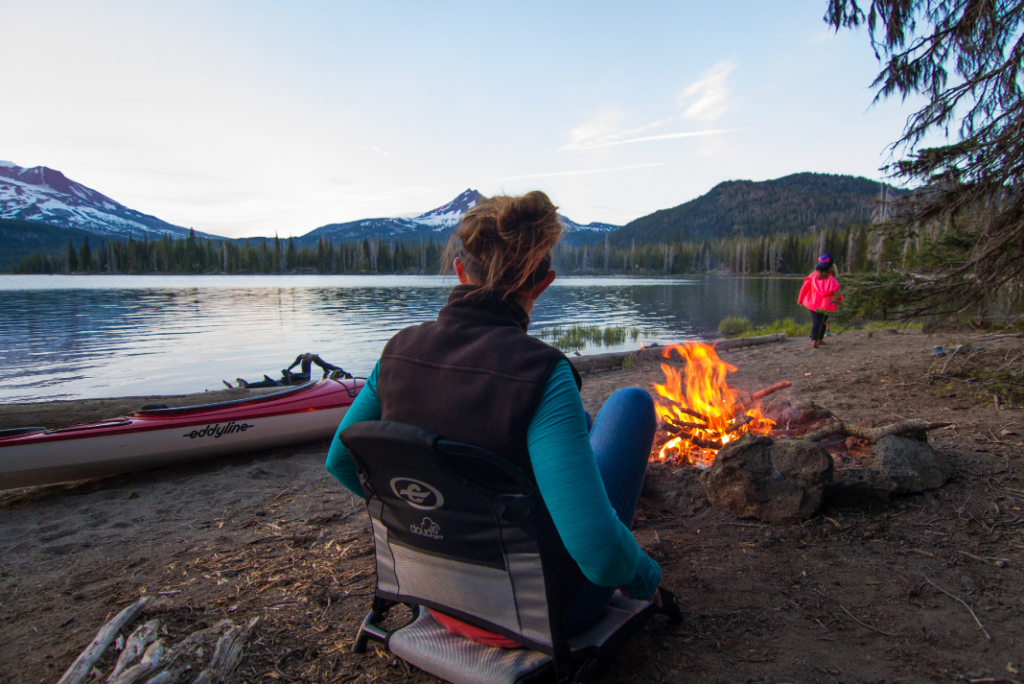 A thoughtful woman sits by the fire and her kayak while looking out to the lake