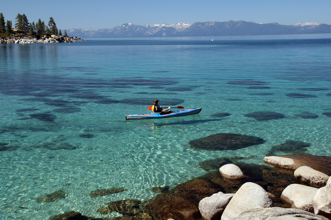Man paddling inflatable kayak on clear waters with mountain and island in background.