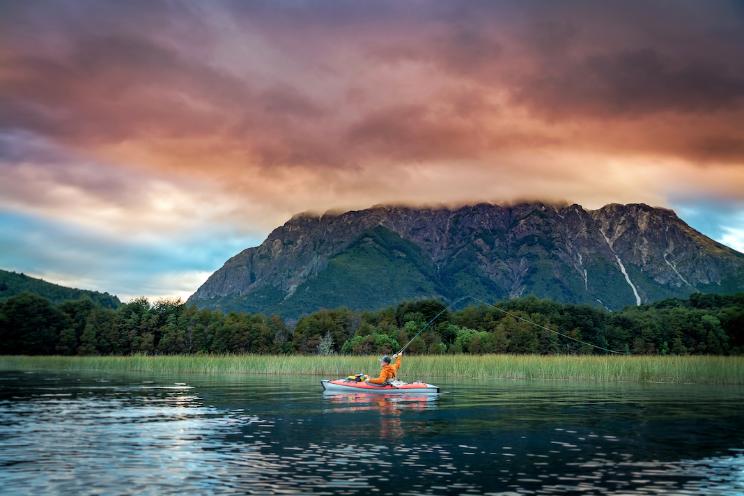 Man casting a line from an inflatable kayak with mountain in background.