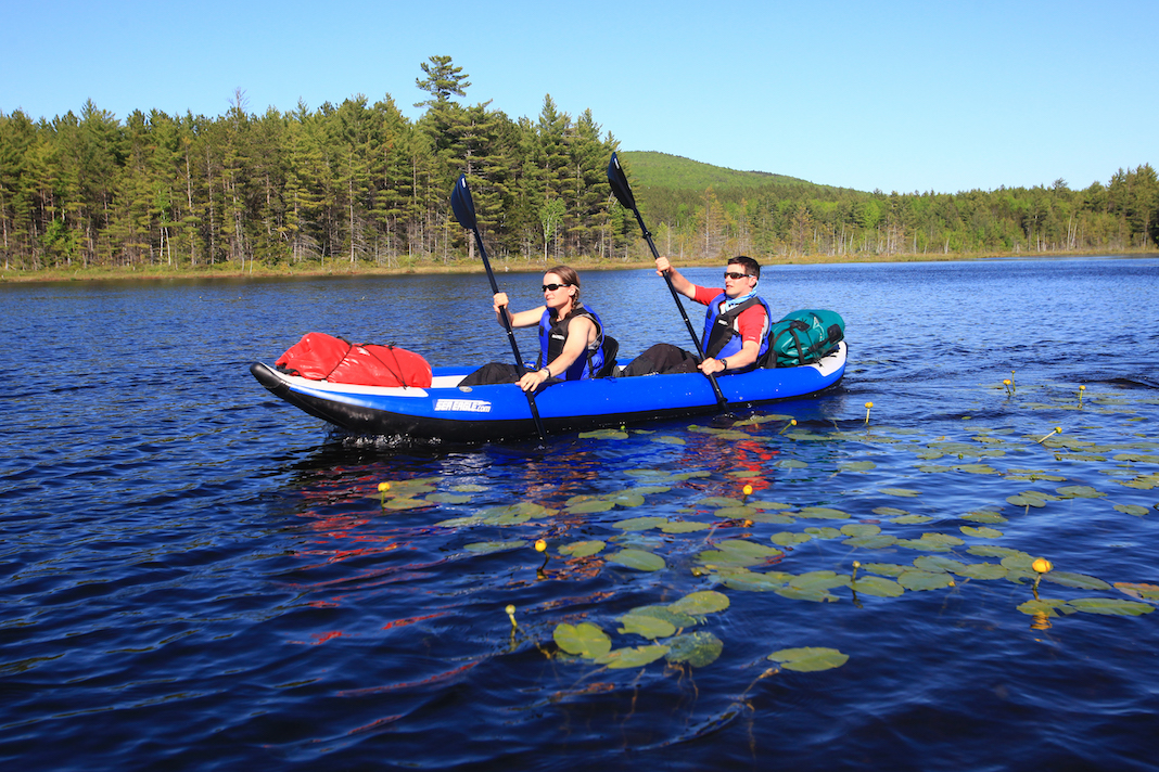 Two people paddling tandem inflatable kayak with bags strapped to stern and bow.