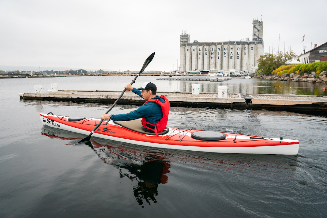 Man paddling red and white touring kayak with factory in background