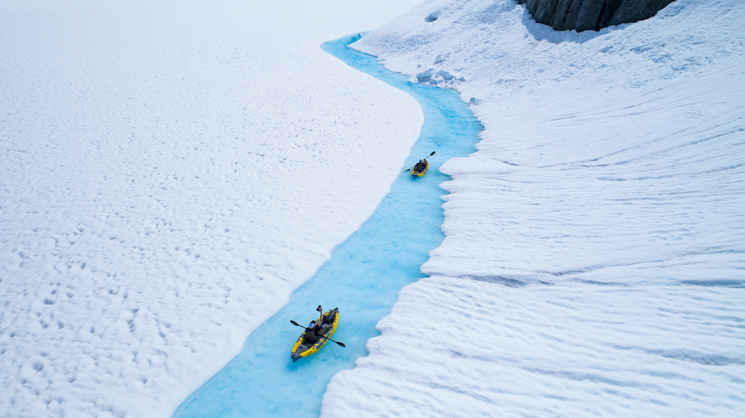 Two kayaks being paddled on narrow rive running between snowy shores