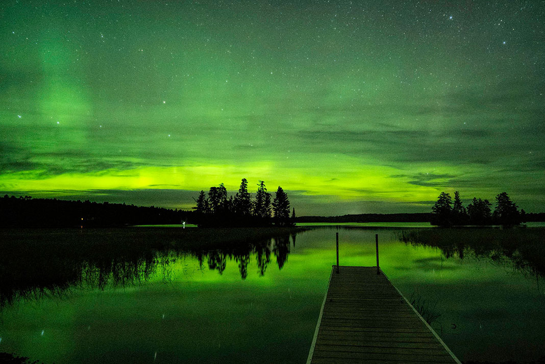 green northern lights appear over lakeside dock