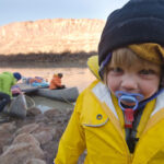 29 Tips For Fun, Worry-Free Family Camping young child with soother and rain coat stands in front of family preparing canoes on a camping trip