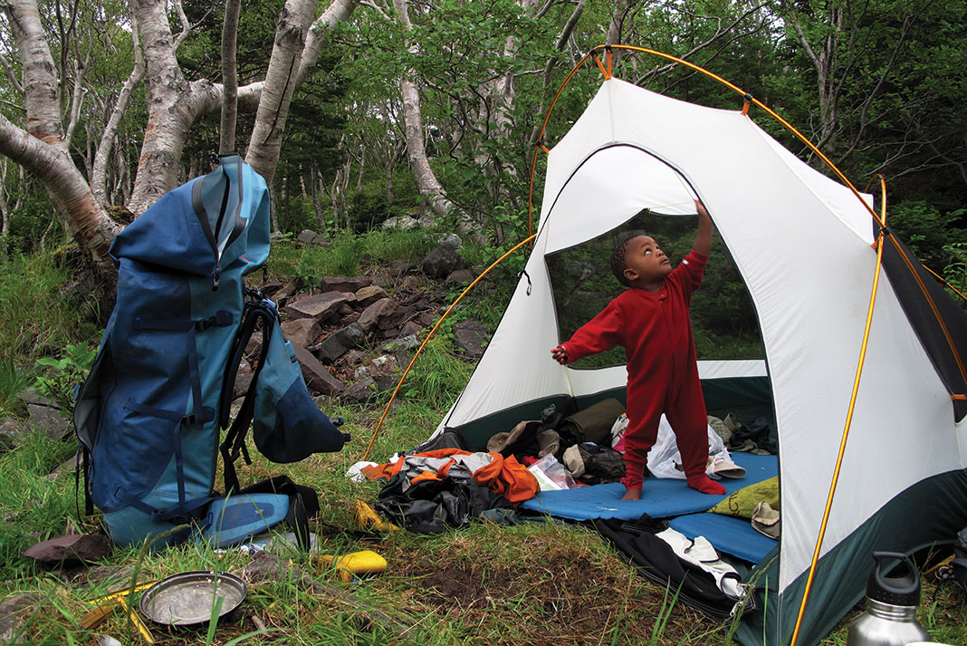 young boy stands up in a tent in the forest with a hiking backpack and gear around him