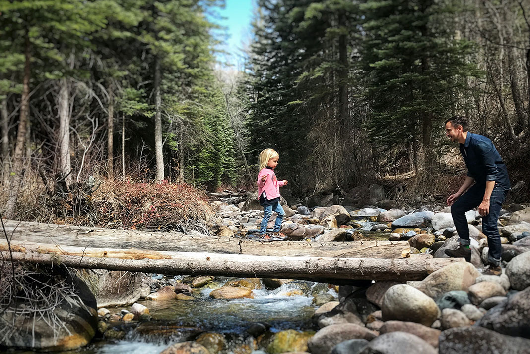young girl crosses a log bridge over a forest brook while her father encourages her