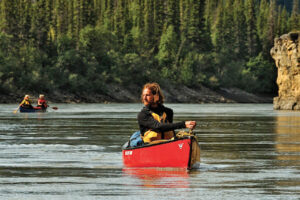 man paddles canoe along a northern river with a second canoe upriver in the background