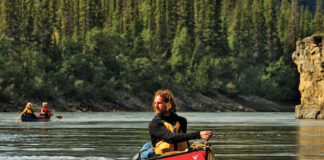 man paddles canoe along a northern river with a second canoe upriver in the background