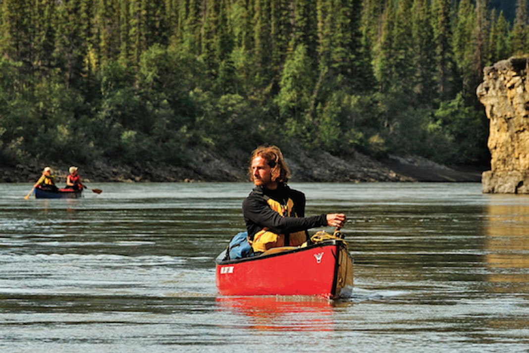 man paddles canoe along a northern river with a second canoe upriver in the background