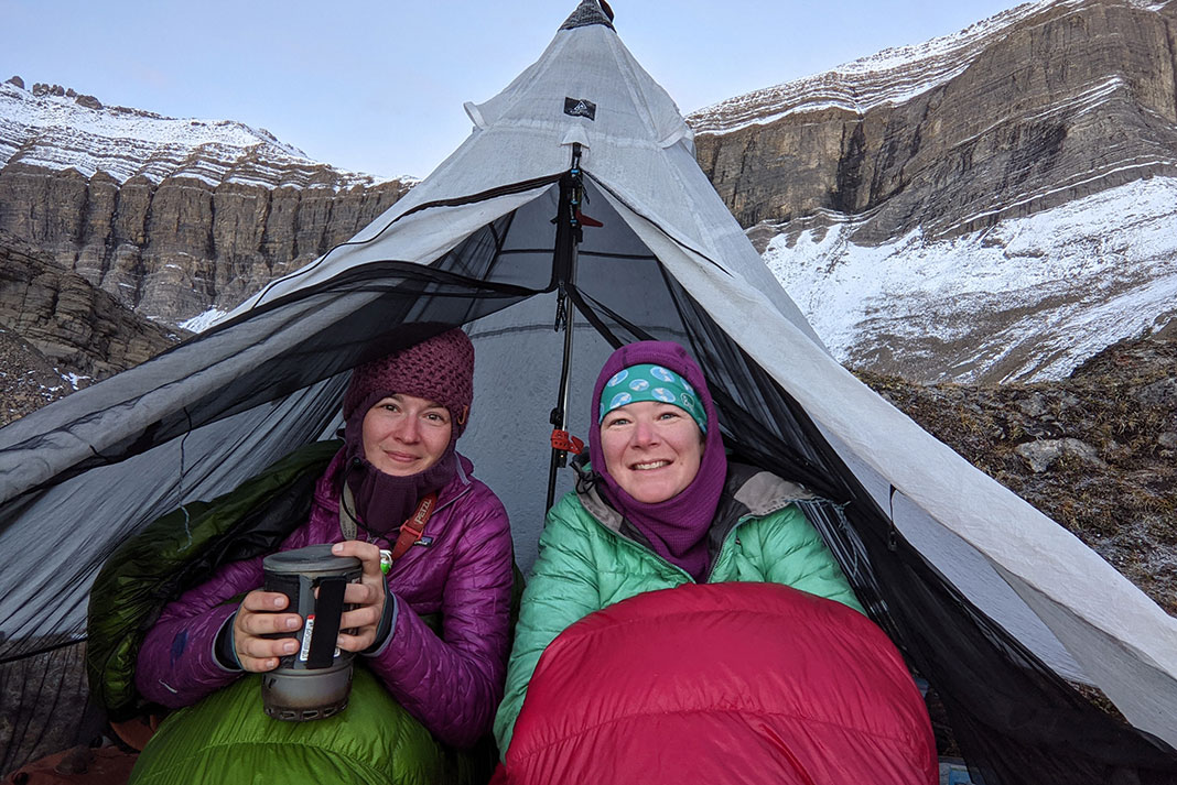 two women sit bundled up in a tent in front of a snowy cliff