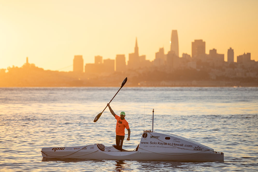 expedition paddler holds up a paddle in front of a city skyline at dawn