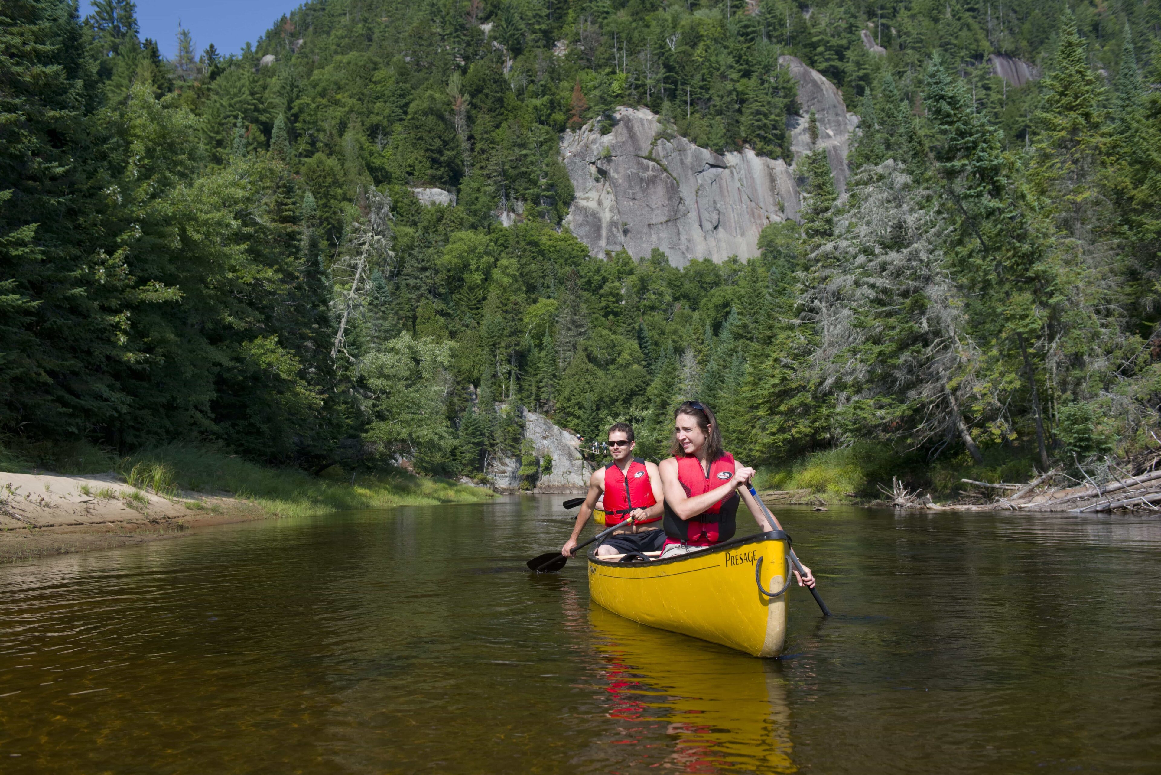 Man and woman paddle yellow canoe on river with cliff in background.