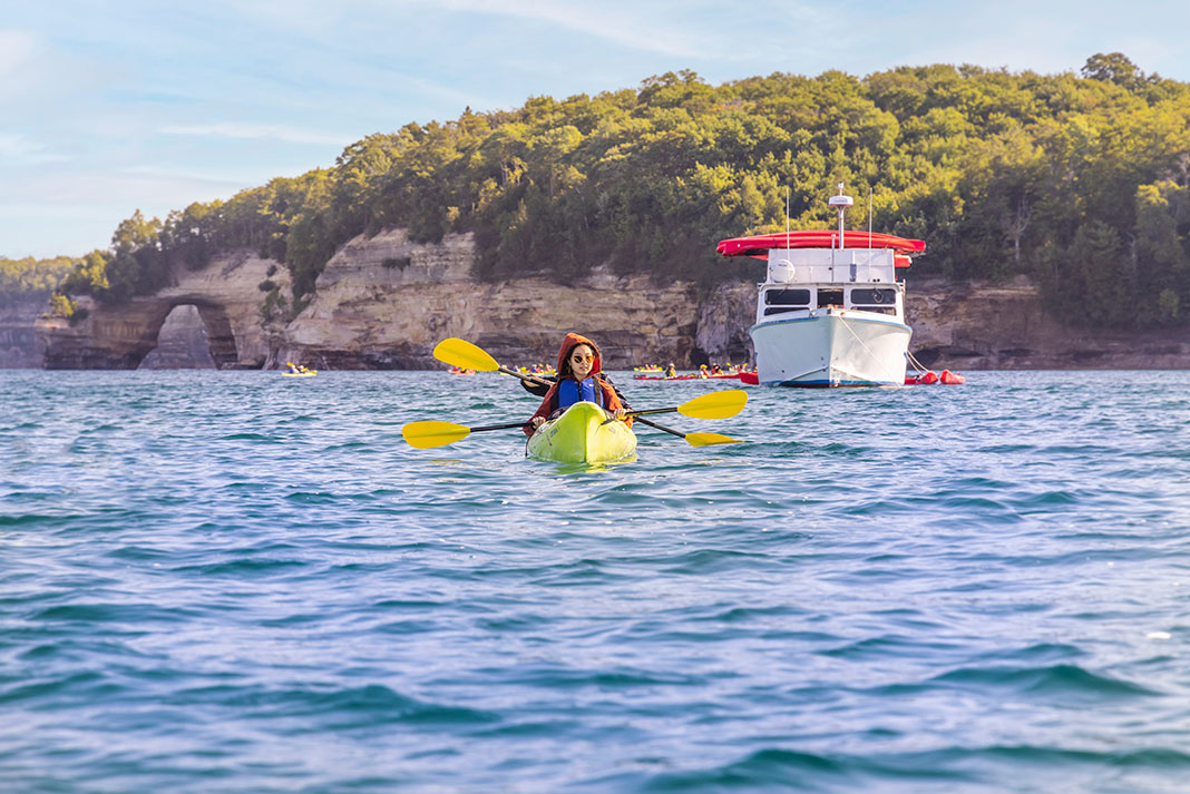 Woman in hooded sweatshirt paddles kayak in front of mothership at Pictured Rocks in Michigan.