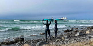 a man and woman in wetsuits stand on a rocky, wave-strewn shore balancing surboards on their heads
